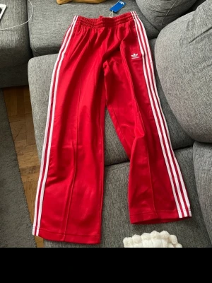 Röda Adidas mjukisbyxor med vita ränder - Snygga röda mjukisbyxor från Adidas med klassiska vita ränder längs sidorna och broderad logga på ena benet. Byxorna har resår i midjan och raka ben. Perfekta för en sportig och avslappnad stil.