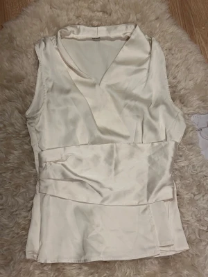 Satintopp - Snygg ärmlös blus i omlottmodell med bred midjedetalj. Tillverkad i ett glansigt, satinliknande material som ger en elegant känsla. Perfekt för sommaren! ‼️Oanvänd
