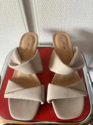 Beige slip-in pumps från Alley - Snygga beige slip-in pumps från Alley med trendig fyrkantig tå och tvinnade remmar i mjukt konstläder. Skorna har en låg klack och öppen häl, perfekta för en stilren look. Passar dig som gillar minimalistisk och modern design.