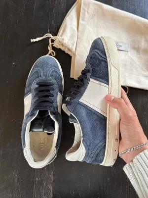 Valentino flycrews - Snygga sneakers från Valentino Garavani i blå mocka och vitt skinn. Klassisk låg modell med svarta snören, vita detaljer och coola nitar på sulan. Sulan har ett unikt mönster undertill. Perfekta för dig som vill ha en stilren men ändå lyxig look. Köptes här på plick i börkan av året. Storlek 38 men passar även 39. Skick mellan 7/10 och 8/10. Ena skon saknar 3 nitar, annars är dom i bra skick, skor och dustbags hänger med. Priset går att diskuteras! Tveka inte att skriva om du har någon fråga!