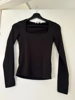 Svart långärmad topp med fyrkantig ringning - En svart långärmad topp med snygg fyrkantig ringning. Modellen är figurnära och passar perfekt till jeans eller kjol. Tillverkad i mjukt och stretchigt material för en bekväm känsla.