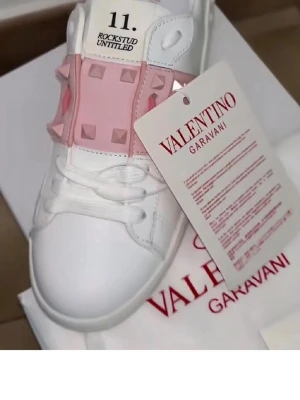 Valentino Rockstud Untitled sneakers - Snygga vita sneakers från Valentino Garavani med rosa band och ikoniska pyramidnitar över snörningen. Skorna har en klassisk låg siluett och är tillverkade i slätt läder. Perfekta för dig som vill sticka ut med exklusiva detaljer och modern design.