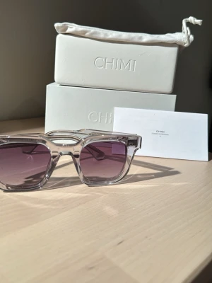 Chimi transparenta solglasögon - Snygga solglasögon från Chimi med transparent båge och fyrkantig form. Glasen är mörka med en lätt toning som går från lila till grått. Perfekta för att lyfta din stil under soliga dagar.