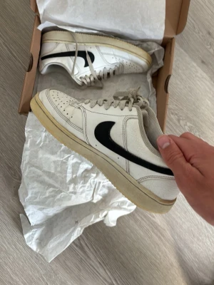 Vita Nike sneakers med svart swoosh - Säljer ett par klassiska vita Nike sneakers med svart swoosh på sidan. Skorna har lågt skaft, perforerad tåbox och beige sula. Tillverkade i läder med textilfoder och snörning framtill. Perfekta för en clean och sportig look.