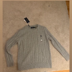 Grå kabelstickad tröja Polo Ralph Lauren - Snygg grå kabelstickad tröja från Polo Ralph Lauren med rund halsringning och lång ärm. Tröjan har det klassiska broderade logotypen på bröstet och ribbade muddar vid ärmslut och nederkant. Perfekt för lager på lager och stilren look.
