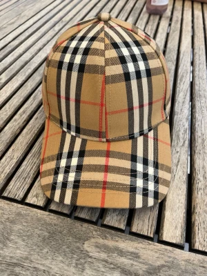 Beige rutig keps från Burberry. Säljer eller byter - Säljer eller byter en klassisk keps från Burberry i beige med det ikoniska rutmönstret i svart, vitt och rött. Kepsen har böjd skärm och justerbar baksida. Tillverkad i bomull för en skön och stilren look.