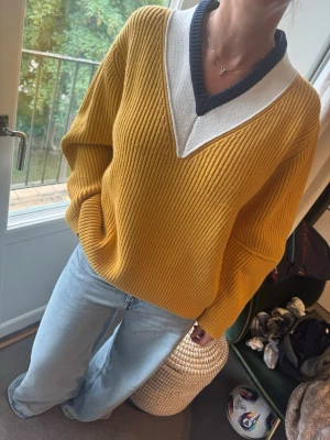 Gul stickad tröja med V-ringning - Trendig gul stickad tröja med bred ribbstickning och djup V-ringning. Kragen är vit med en svart kant som ger en sportig känsla. Tröjan har avslappnad passform och långa ärmar. Perfekt för dig som gillar en färgklick i garderoben. (Pris kan diskuteras)