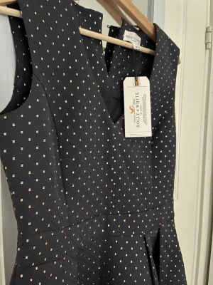 Svart klänning Holly & Whyte - Svart klänning från Holly & Whyte med små guldiga prickar. Ärmlös modell med markerad midja och lätt utställd kjol. Klänningen har en elegant och klassisk siluett, perfekt för dig som gillar stilrena detaljer och diskret mönster.