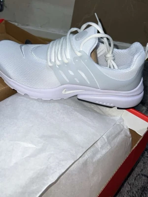 Nike vita mesh sneakers - Snygga vita sneakers från Nike med mesh-ovandel och plastdetaljer på sidorna. Skorna har en låg profil, vita skosnören och en bekväm, vadderad insida. Perfekta för dig som gillar stilrena och sportiga skor.