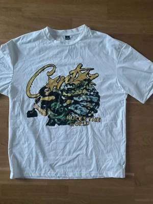 Vit t-shirt med tryck från CMTX - Snygg vit t-shirt från CMTX med stort grafiskt tryck i gult, svart och grönt på framsidan där det står 'CMTX RULES THE WORLD'. Klassisk passform och rund hals. Perfekt för dig som gillar streetwear och statement-plagg.