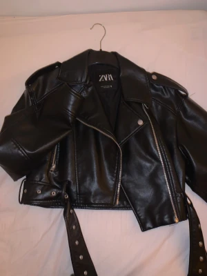 Svart skinnjacka från Zara - Snygg svart skinnjacka från Zara med klassisk bikerstil, breda slag, silverfärgade dragkedjor och knappar samt bälte i midjan. Kortare modell som ger en cool och trendig look. Perfekt att matcha med jeans eller klänning.