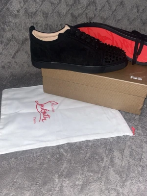 Svarta Lou Spikes sneakers Louboutin - Säljer ett par svarta Lou Spikes sneakers från Christian Louboutin. Skorna är i mocka med svarta nitar på tån och har den ikoniska röda sulan. Snygg och edgy design med svarta skosnören och låg profil. Perfekta för dig som vill sticka ut med exklusiva detaljer.