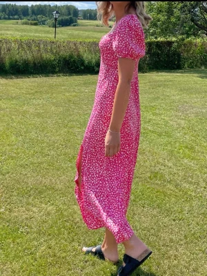 Rosa blommig långklänning med slits - Söt rosa långklänning med vitt blommönster och puffärmar. Klänningen har en smickrande v-ringning och en hög slits på sidan som ger en trendig look. Perfekt för varma dagar och sommarens alla tillfällen.