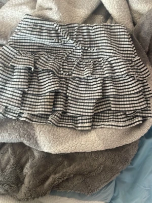 Randig volangkjol med svartvita ränder - Kort kjol med svartvita ränder och flera volanglager. Kjolen har en elastisk midja och ett lekfullt, rynkat fall. Perfekt för dig som gillar en trendig och ungdomlig stil.