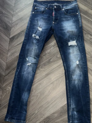 Dsquared2 jeans med slitningar - Säljer ett par blå jeans från Dsquared2 med slitningar och trasiga detaljer på framsidan. Jeansen har fem fickor, knappgylf och en smal passform. Materialet är denim med en cool tvätt och distressed look storlek size 30 så de e ungefär som xs tveka hör av dig om du undrar något eller vill ha fler bilder, pris går att diskutera