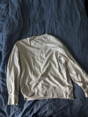 Beige sweatshirt från Uniqlo - En stilren beige sweatshirt från Uniqlo i mjuk bomull. Tröjan har rund halsringning, ribbade muddar och en klassisk V-detalj vid halsen. Perfekt för en avslappnad och enkel look.