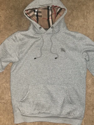 Grå hoodie från Burberry med huva - Snygg grå hoodie från Burberry med klassiskt rutigt mönster i huvan och broderad logga på bröstet. Hoodien har dragsko i huvan och en stor magficka. Perfekt för dig som gillar stilrena plagg med exklusiva detaljer.
