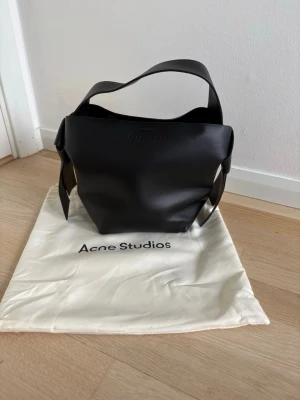 Svart handväska Acne Studios - Snygg svart handväska från Acne Studios i slätt läder. Väskan har en minimalistisk design med bred axelrem och präglad logga framtill. Perfekt för dig som gillar stilrena accessoarer och vill ha en väska som sticker ut utan att vara för mycket.