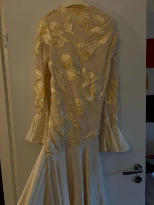 Lång champagnefärgad klänning med spets - Elegant långklänning i champagnefärg med stora blommiga spetsapplikationer och vida långa ärmar. Klänningen har en mjuk, flowy kjol och detaljerade broderier över hela överdelen. Perfekt för dig som vill sticka ut med en unik och romantisk look.