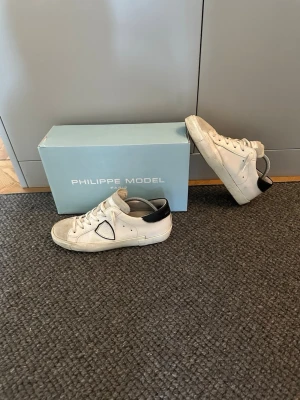 Vita Philippe Model sneakers - Snygga vita sneakers från Philippe Model med klassisk låg siluett. Skorna har detaljer i svart på hälen, vita skosnören och en ikonisk logga på sidan. Tillverkade i läder och mocka för en stilren och trendig look. 