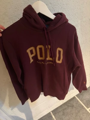 Vinröd hoodie från Polo Ralph Lauren - Säljer en vinröd hoodie från Polo Ralph Lauren med stor POLO-logga i gult på bröstet. Hoodien har huva med dragsko och är tillverkad i mjukt bomullsmaterial. Perfekt för en chill och stilren look.