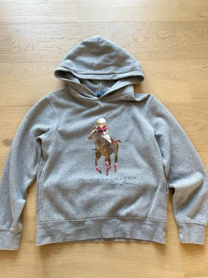 Grå hoodie Polo Ralph Lauren M - Säljer en grå hoodie från Polo Ralph Lauren med ikoniskt Polo Bear-motiv på bröstet. Hoodien har huva och ribbade muddar vid ärmar och nederkant. Tillverkad i mjukt bomullsmaterial och perfekt för en avslappnad stil.