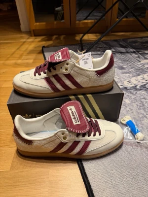 Adidas Wales Bonner sneakers vinröd/vit - Snygga Adidas Wales Bonner sneakers i vitt och vinrött med klassiska tre ränder på sidan. Ovandelen är i mocka och läder, med detaljer i guld och gummisula. Tungan har Wales Bonner-logga och skorna har en retro vibe. Perfekta för dig som gillar unik stil.