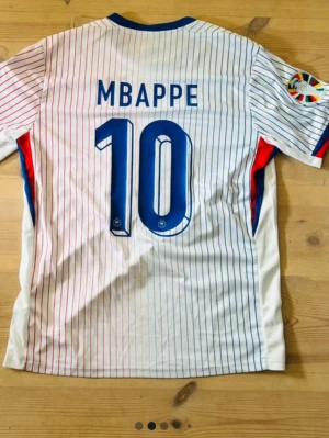 Frankrike Mbappé matchtröja Nike - Säljer en vit matchtröja från Nike med tunna blå och röda ränder, Frankrikes landslagsmärke och nummer 10 Mbappé på ryggen. Tröjan har blå detaljer vid krage och ärmslut samt broderad Nike-logga. Perfekt för fotbollsfans!