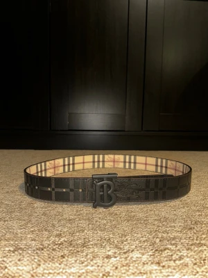 Burberry vändbart bälte  - Snyggt vändbart bälte från Burberry med klassiskt rutigt mönster på ena sidan i beige, rött och svart samt svart rutigt mönster på andra sidan. Stor B-logga i metall framtill. Perfekt accessoar för att lyfta din outfit. Bältet är 106cm