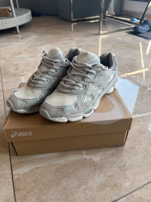 Asics Gel Nyc Cream/Cloud Grey - Snygga beige sneakers från Asics med ovandel i cream och cloud grej. Skorna har klassisk snörning, vadderad plös och häl samt detaljer i olika nyanser av beige och grått. Perfekta för en sportig och trendig look i storlek 44