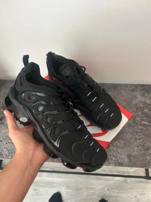Nike Air Vapormax Plus svarta sneakers - Nike Air Vapormax Plus i helsvart med en chunky sula och synliga luftbubblor. Ovandelen är i mesh och syntet med vågiga detaljer och klassisk Nike-logga på hälen. Grymt futuristisk look och riktigt sköna att gå i.