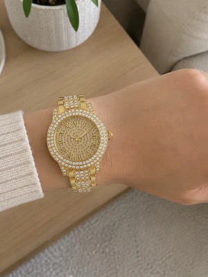 Guldig klocka med stenar och romerska siffror - Snygg guldig klocka med glittrande stenar runt urtavlan och på armbandet. Urtavlan har romerska siffror och ett detaljerat mönster. Perfekt accessoar för dig som vill sticka ut och addera lite extra bling till din stil.