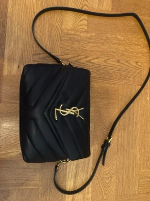 Svart YSL axelväska i läder - Säljer en svart axelväska från Saint Laurent med klassisk YSL-logga i guld på framsidan. Väskan är quiltad i mjukt läder och har justerbar axelrem med gulddetaljer. Perfekt storlek för det viktigaste och ger en lyxig känsla till din outfit.