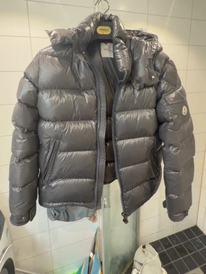 Mörkgrå dunjacka från Moncler - Snygg mörkgrå dunjacka från Moncler med glansig finish och klassisk puffer-design. Jackan har hög krage, avtagbar huva och två sidofickor med dragkedja. Moncler-logga på ärmen och justerbara muddar. Perfekt för kalla vinterdagar.