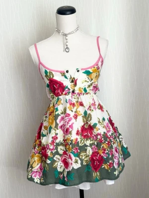 Blommig sommarklänning med knappar - Söt blommig klänning med smala axelband och knappar framtill. Klänningen har en vit bas med färgglada blommor i rosa, rött, gult och grönt samt rosa kantband. Perfekt för varma dagar och ger en romantisk vibe.