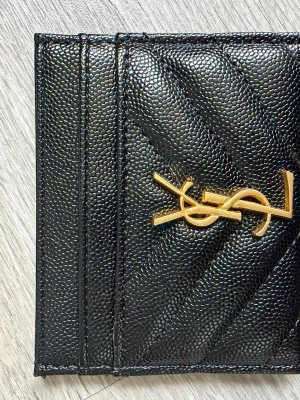 Svart YSL korthållare i läder - Snygg svart korthållare från Saint Laurent i präglat läder med guldfärgad YSL-logga framtill. Klassisk och stilren design med flera kortfack och chevronmönster. Perfekt accessoar för dig som gillar lyxiga detaljer och tidlös stil.