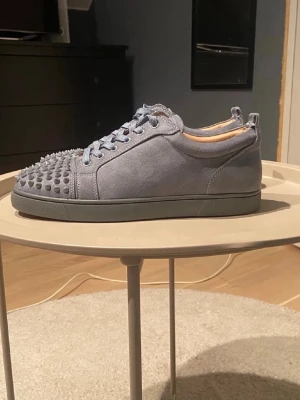 Gråa Christian Louboutin sneakers - Snygga gråa sneakers från Christian Louboutin i mocka med ikoniska röda sulor och coola nitar på tån. Skorna har lågt skaft, matchande grå skosnören och en unik design som sticker ut. Perfekta för dig som vill ha något extra till din outfit.
