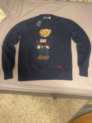 Marinblå stickad tröja Polo Bear - Marinblå stickad tröja från Polo Ralph Lauren med den ikoniska Polo Bear på bröstet och RL-broderi i rött nedtill. Tröjan har rund halsringning, ribbade muddar och är långärmad. Perfekt för dig som vill ha en klassisk och lekfull look.