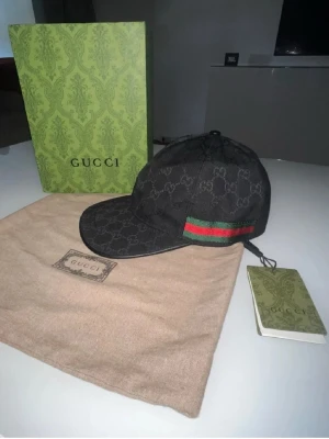 Gucci keps  - Snygg svart keps från Gucci med det klassiska GG-monogrammet över hela kepsen. På sidan finns en grön och röd rand som ger en ikonisk touch. Kepsen har böjd skärm och justerbar rem bak. Materialet är troligtvis bomull eller canvas, kepsen är helt ny och är öppen för förslag!