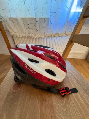 Röd och vit cykelhjälm från Hamax - Snygg cykelhjälm i rött och vitt från Hamax med justerbart spänne och flera ventilationshål. Hjälmen har en sportig design och är tillverkad i lätt plast och skum för bra komfort och skydd.