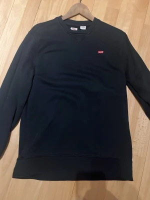 Svart sweatshirt från Levi's - Klassisk svart sweatshirt från Levi's med rund halsringning och diskret röd Levi's-logga på bröstet. Tröjan har långa ärmar och ribbade muddar vid ärmslut och nederkant. Perfekt för en avslappnad och stilren look.