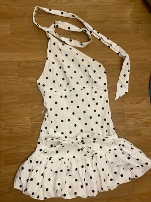 Vit prickig oneshoulder klänning - Säljer en vit kortklänning med svarta prickar och oneshoulder-design. Klänningen har volang nedtill och knytband vid axeln för en snygg detalj. Perfekt för dig som vill sticka ut med en lekfull och trendig look.