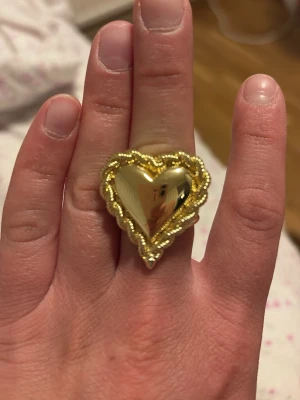 Guldfärgad hjärtformad ring - Säljer en statementring i guldfärg med ett stort hjärta som motiv. Ringen har en kant med små pärlliknande detaljer runt hjärtat som ger en lyxig känsla. Perfekt accessoar för dig som vill sticka ut och addera lite extra bling till din stil.