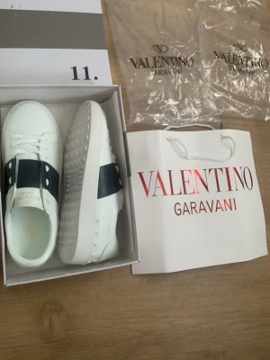 Valentino Garavani vita sneakers - Snygga vita sneakers från Valentino Garavani med bred svart rem över foten och dekorativa nitar. Skorna har en chunky sula och är tillverkade i slätt läder. Perfekta för dig som vill ha en exklusiv och trendig look.