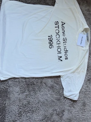 Acne Studios t-shirt vit  - Vit t-shirt från Acne Studios med svart tryck framtill där det står 'Acne Studios STOCKHOLM 1996'. Klassisk passform och rund hals. Tillverkad i mjuk bomull, perfekt för en clean och stilren look.