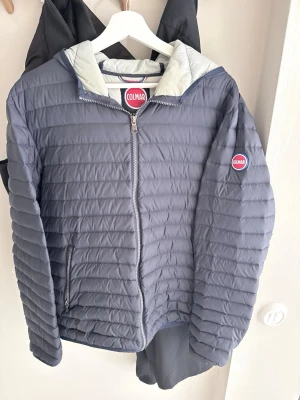 Colmar jacka - Snygg blå pufferjacka från Colmar med quiltad design och huva. Jackan har dragkedja framtill, två sidofickor med dragkedja och Colmar-logga på ärmen. Insidan är ljusgrå och materialet är lätt och värmande, perfekt för kalla vinterdagar.