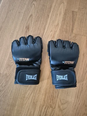 Svarta Everlast MMA-handskar - Säljer ett par svarta MMA-handskar från Everlast med öppen fingertopp och bred kardborrestängning. Handskarna har TITAN-logga på ovansidan och är tillverkade i syntetmaterial. Perfekta för träning och kampsport.