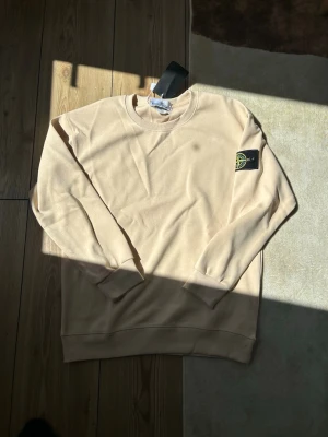 Beige sweatshirt från Stone Island - Snygg beige sweatshirt från Stone Island med klassisk rund hals och ribbade muddar. Ikonisk svart patch med logotyp på vänster ärm. Perfekt för dig som gillar stilrena och trendiga plagg. Materialet är mjukt och bekvämt, passar till många outfits.