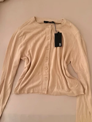 Beige kofta från 157 - Stilren beige kofta från 157 med rund halsringning och knappar framtill. Långärmad modell i mjukt, tunt material som passar perfekt till lager på lager. Enkel och tidlös design som är lätt att matcha med olika outfits.