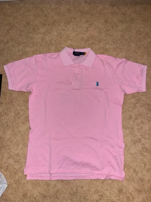 Rosa pikétröja från Polo Ralph Lauren - Säljer en klassisk rosa pikétröja från Polo Ralph Lauren med blå logga broderad på bröstet. Tröjan har korta ärmar, krage och knappar framtill. Perfekt för dig som gillar stilrena och färgglada plagg. Storlek XL fast den är liten i storlek så passar dig som har L bättre. Helt ny och oanvänd så pris kan diskuteras vid snabb affär!